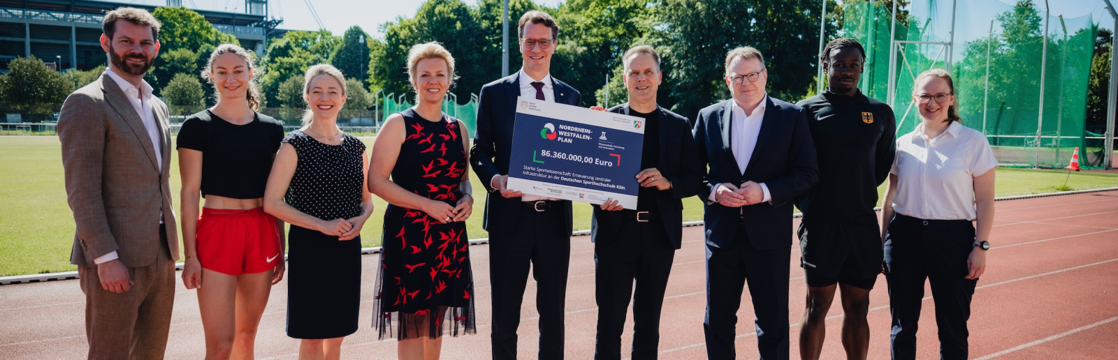 Ministerpräsident Wüst umgeben acht Personen auf einer Tartanbahn in einem Stadion, er hält ein Schild in der hand
