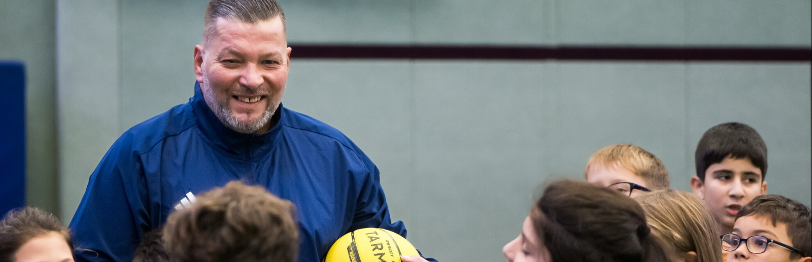 Mann in blauer Sportkleidung hält einen gelben Ball, er ist umringt von Jungen und Mädchen 
