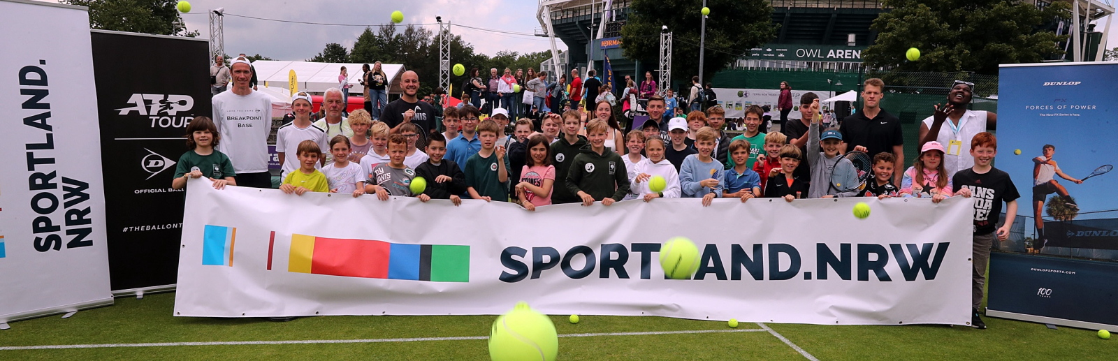 Auf einem Rasen-Tenniscourt bei den Terra Wortmann Open sind Kinder hinter einem Sportland.NRW-Banner und werfen gelbe Bälle