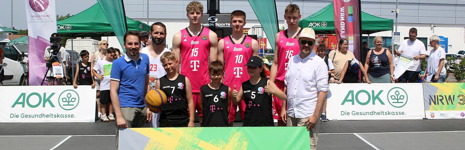 Gruppenfoto beim Basketball-Event „NRW 3x3 Tour“ mit Spielern in rosa-schwarzen Trikots, im Hintergrund sind Banner und Zuschauer zu sehen.