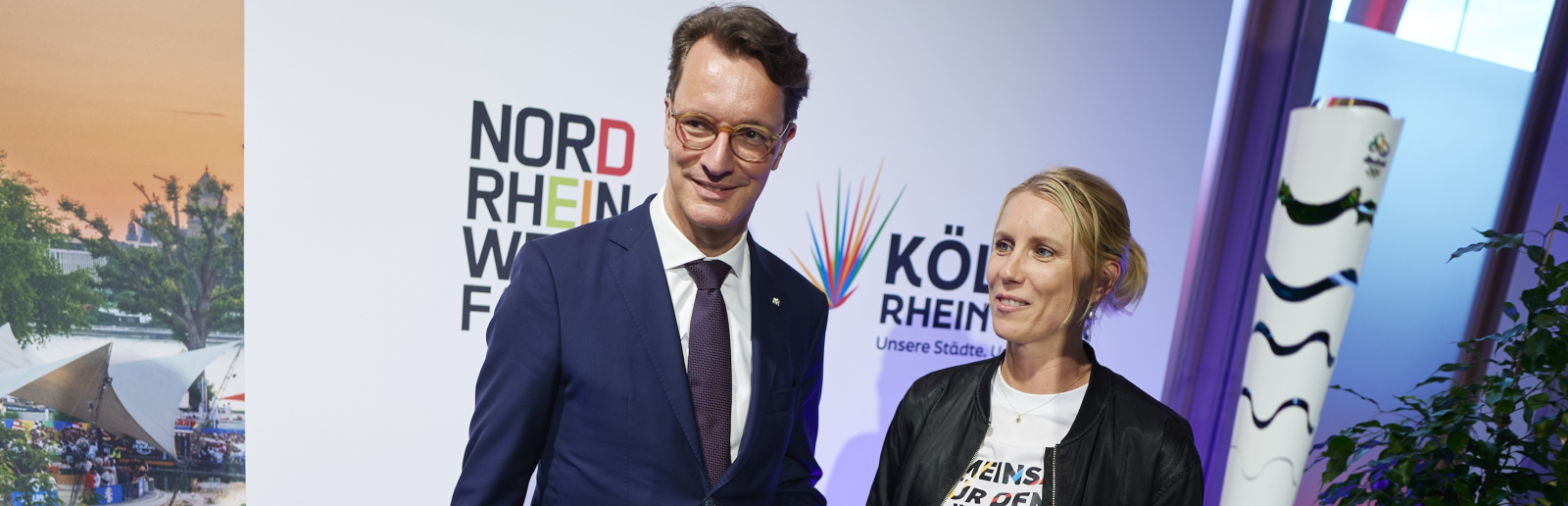 Hendrik Wüst neben einer Frau und der Olympiafackel von Rio de Janeiro auf einem Podest, beide stehen vor einer Wand mit dem Schriftzug der Bewerbung KölnRheinRuhrn