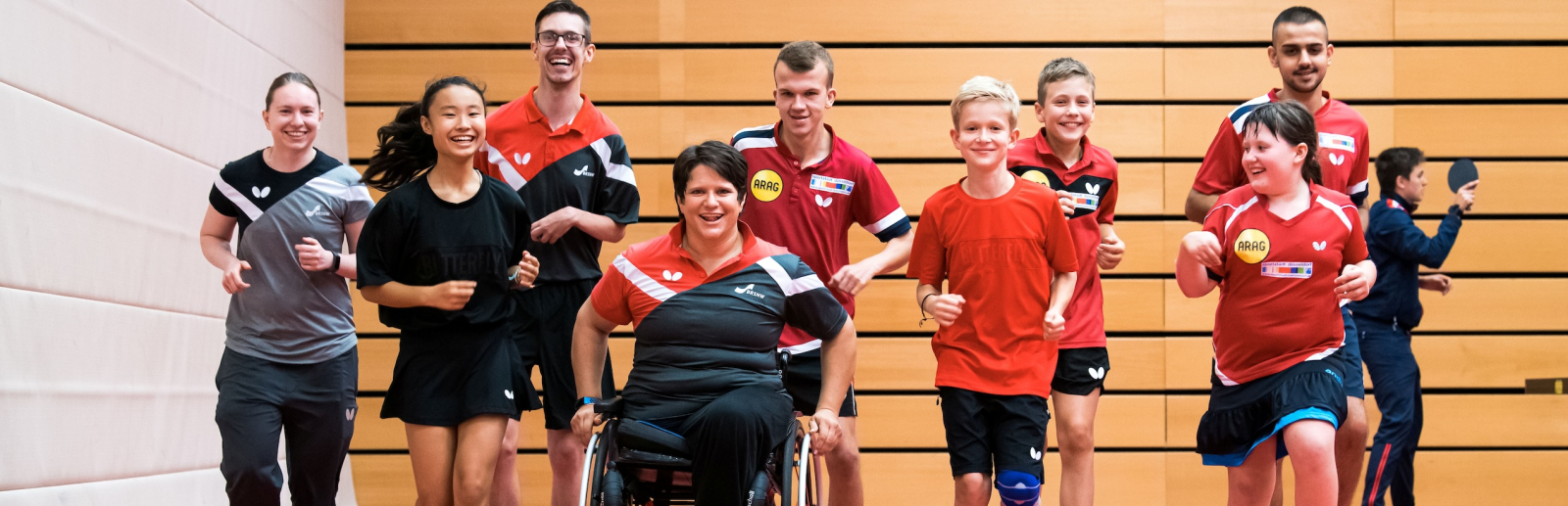 Gruppe von Sportlerinnen und Sportlern in einer Halle mit rotem Boden, eine davon in eiem Rollstuhl