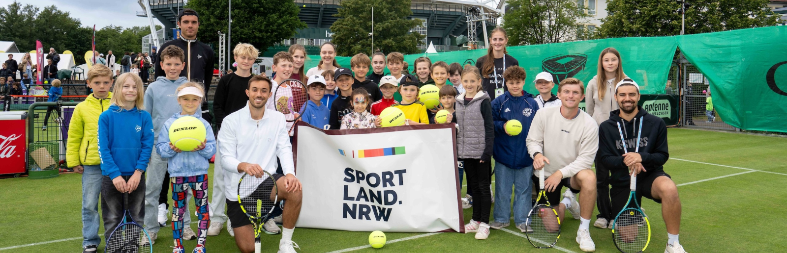 Gruppe von Kindern halten ein Banner mit "Sportland.NRW" auf einem Rasen-Tennisplatz