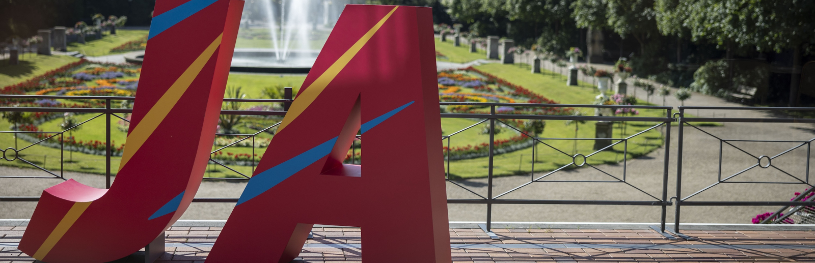 Große Buchstaben J und A in rot vor einem Park mit Blumen und Bäumen