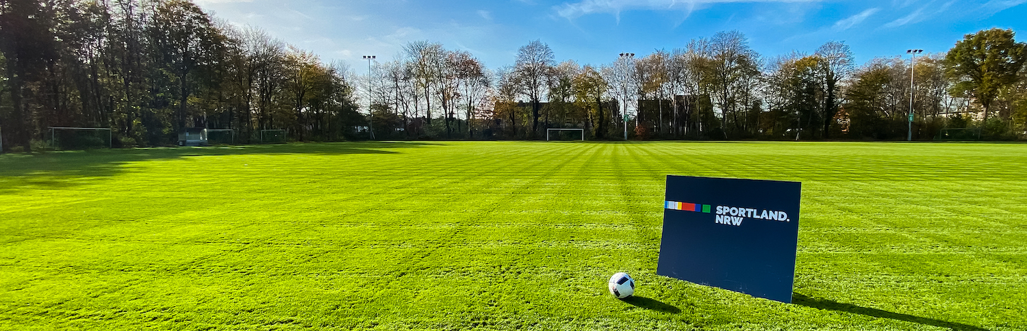 Grüne Rasenfläche, umgeben von Bäumen, auf der ein Sportland.NRW-Schild steht, danaeben liegt ein Fußballball