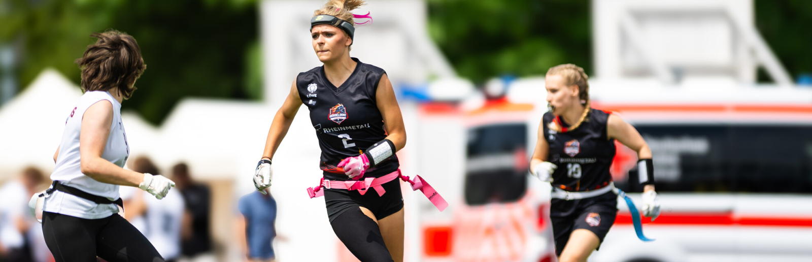 Junge Frau in schwarzer Sportkleidung spielt Flag Football auf einer Wiese, im Hintergrund stehen Fahrzeuge