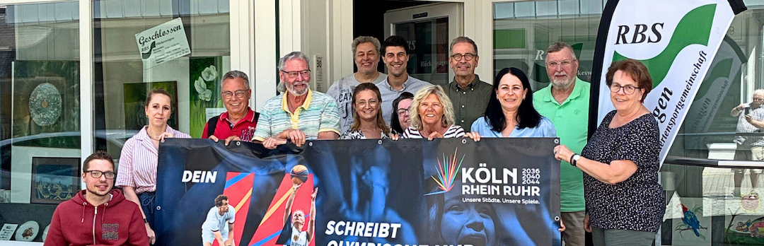 Gruppe von Menschen, davon einer in einem Rollstuhl, vor einem Gebäude. Sie halten ein Banner der Bewerbung KölnRheinRuhr