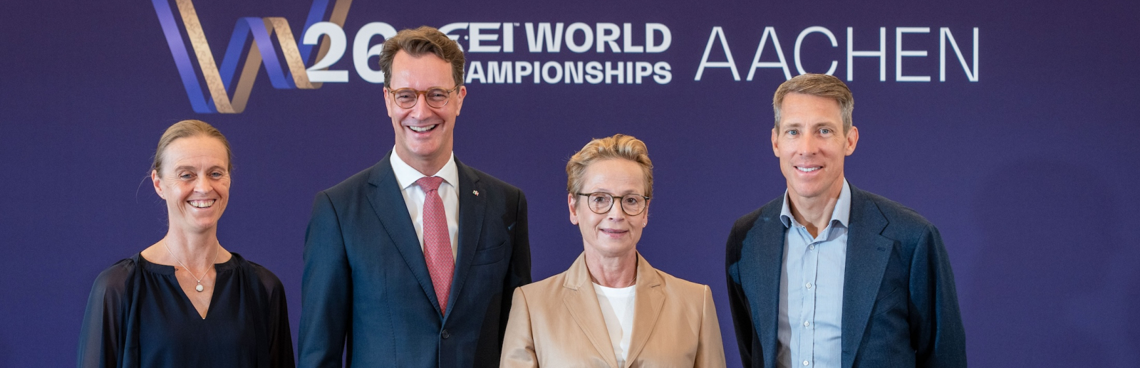 Ministerpräsident Wüst und drei Pferdesport-Athletinnen und -Athleten vor einem Banner der Reit-WM in Aachen, vor ihnen ein goldfarbenes Schachfigur-Pferd