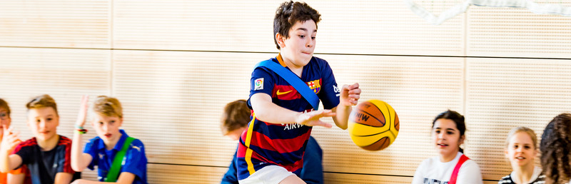 Schulikinder auf einer Bank in einer Turnhalle, davor ein Junge mit einem Basketball