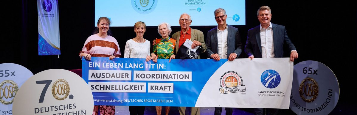 Sechs Personen halten einen Banner bei einer Veranstaltung, wo der 100-jährige Johann Otte als ältester Teilnehmer beim Deutschen Sportabzeichen ausgezeichnet wurde.