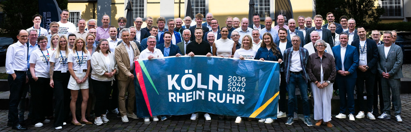 Gruppe von Menschen vor einem beigefarbenen Altbau mit einem Banner der Bewerbung KRR