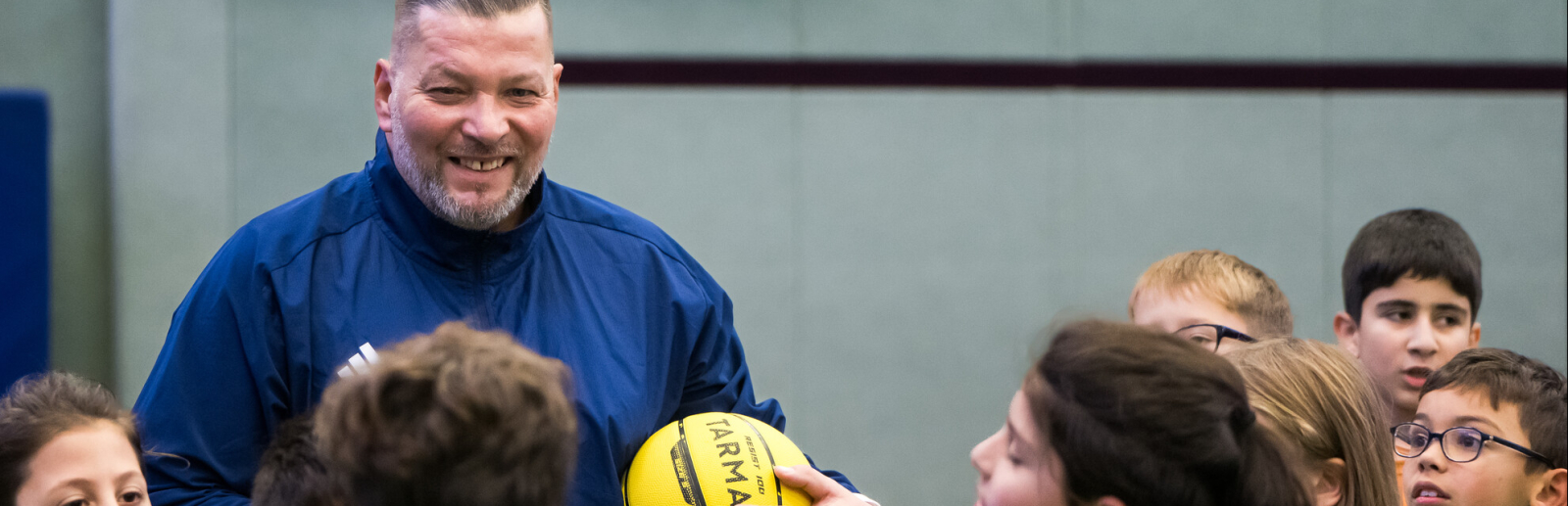 Mann mit blauem Triikot hält einen gelben Ball,, vor ihm Kinder in einem Kreis