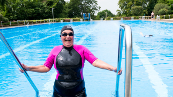 Eine Schwimmerin im pink-schwarzen Neoprenanzug, mit schwarzer Badekappe und schwarzer Schwimmbrille steigt aus einem offenen Schwimmbecken, ihre beiden Hände jeweils links und rechts am Geländer.