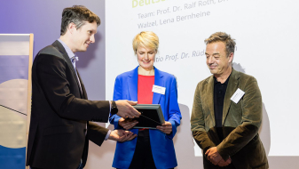 Drei Personen, in der Mitte eine Frau, bei der Übergabe einer Urkunde