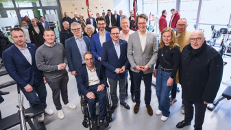 Hendrick Wüst inmitten mehrerer Personen, davon einer im Rollstuhl, in einem Raum mit Fenstern im Hintergrund