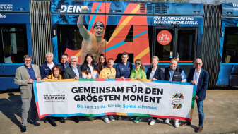 Andrea Milz und weitere Personen mit einem Banner der KRR-Bewerbung vor einer Straßenbahn im "Olympia-Look"