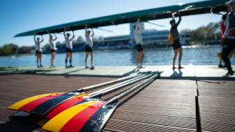 Ruderer mit dem Boot über dem Kopf, im Vordergrund Ruderblätter in den Deutschland-Farben