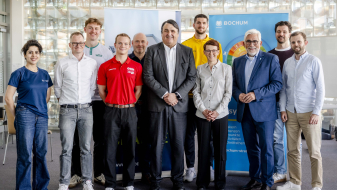 Elf Personen in einem Halbkreis vor zwei Bannern, hinter ihnen eine Trennwand aus Glasbausteinen