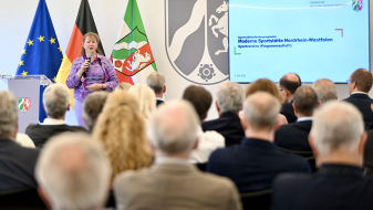Andrea Milz vor drei Fahnen und Videobildschirm, sie spricht zu Teilnehmenden einer Pressekonferenz
