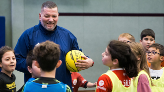 Mann in blauer Sportkleidung hält einen gelben Ball, er ist umringt von Jungen und Mädchen 