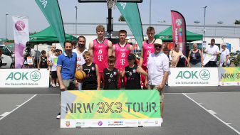 Gruppenfoto beim Basketball-Event „NRW 3x3 Tour“ mit Spielern in rosa-schwarzen Trikots, im Hintergrund sind Banner und Zuschauer zu sehen.