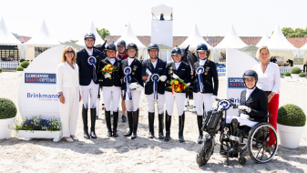 Team von Para Dressurreiterinnen und -reitern, eine davon in einem Rollstuhl, vor einem Hindernisparcours