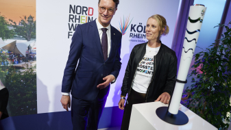 Hendrik Wüst neben einer Frau und der Olympiafackel von Rio de Janeiro auf einem Podest, beide stehen vor einer Wand mit dem Schriftzug der Bewerbung KölnRheinRuhrn