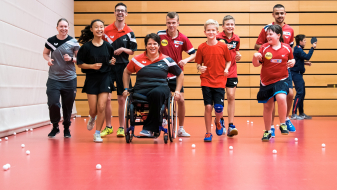 Gruppe von Sportlerinnen und Sportlern in einer Halle mit rotem Boden, eine davon in eiem Rollstuhl