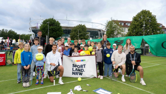 Gruppe von Kindern halten ein Banner mit "Sportland.NRW" auf einem Rasen-Tennisplatz