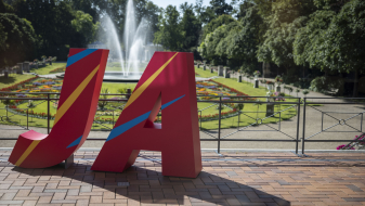 Große Buchstaben J und A in rot vor einem Park mit Blumen und Bäumen