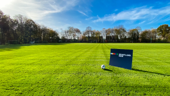 Grüne Rasenfläche, umgeben von Bäumen, auf der ein Sportland.NRW-Schild steht, danaeben liegt ein Fußballball