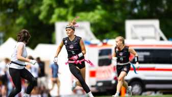 Junge Frau in schwarzer Sportkleidung spielt Flag Football auf einer Wiese, im Hintergrund stehen Fahrzeuge