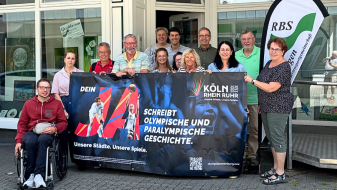 Gruppe von Menschen, davon einer in einem Rollstuhl, vor einem Gebäude. Sie halten ein Banner der Bewerbung KölnRheinRuhr