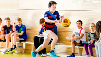 Schulikinder auf einer Bank in einer Turnhalle, davor ein Junge mit einem Basketball