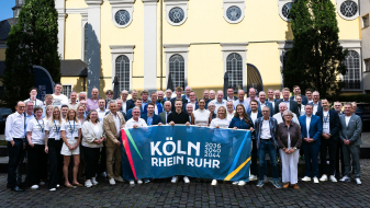 Gruppe von Menschen vor einem beigefarbenen Altbau mit einem Banner der Bewerbung KRR